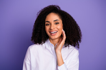 Attractive young woman with curly hair smiling, dressed in white shirt, posing against purple...