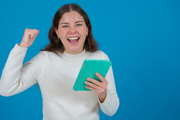 Happy young woman in a white sweater raising her fist in celebration while holding a green tablet and smiling at the camera, against a blue background, symbolizing success, joy, and digital life. 
