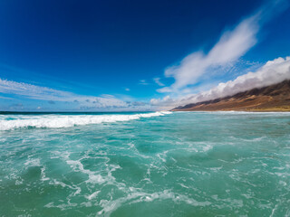 Turquoise waves lap against the sandy beach of Fuerteventura under a clear blue sky, creating a serene coastal atmosphere.
