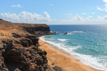 Bright sunlight highlights the sandy beach bordered by rocky cliffs and the gentle waves of the...
