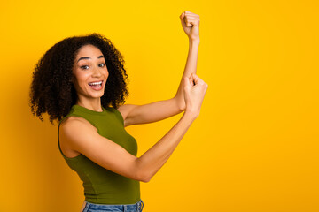 Cheerful young woman in a green top raising fists in a confident pose against a vibrant yellow...