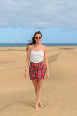 A woman in a strapless top and plaid skirt walks barefoot across golden sand dunes near the ocean...