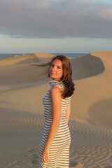 A woman stands gracefully in a striped dress, gazing back while surrounded by soft sand dunes under...