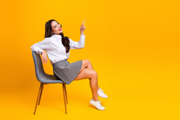 Young woman dressed in formal business attire sits cheerfully on a chair against a vibrant yellow...