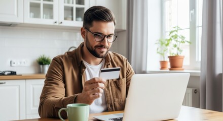 A man with glasses and a beard uses a credit card with his laptop at a desk, engaging in online shopping