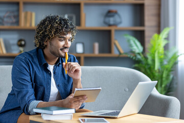 A young man works from home, taking notes while looking at his laptop screen. He appears thoughtful.