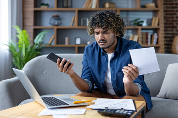 A concerned man reviews bills while holding a phone, working from home and checking finances.