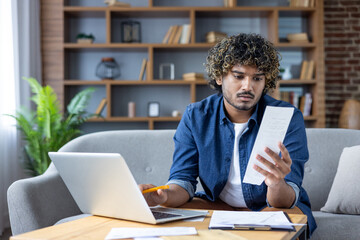 A man with curly hair reviews a receipt while working on a laptop at home.