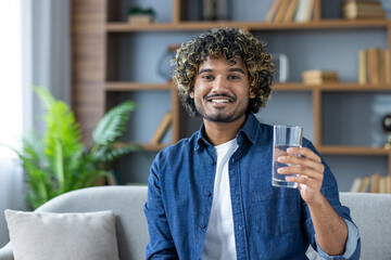 A smiling man holds a glass of water, sitting on a sofa indoors. He appears healthy and happy.