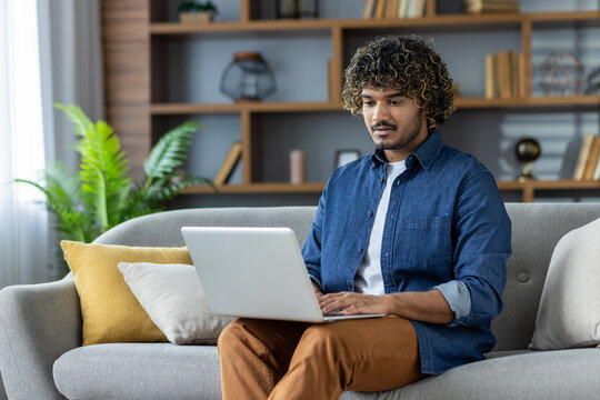 A man works remotely from his home on a laptop, sitting on a sofa. The scene has a cozy and productive feel.