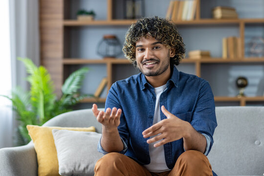 A man is sitting on a couch in his home, talking on a video call while looking at the camera
