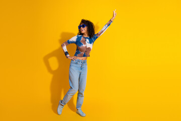 Cheerful young woman posing stylishly in vibrant top and jeans against a bold yellow background,...
