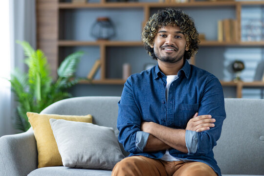 A smiling man with curly hair sits on a couch, arms crossed, wearing a denim shirt in a well-lit living room setting.