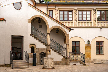 Historic Building At Lenbachsplatz In Munich: Elegant Facade With Decorative Gables, Stone Archway, And Red Roof Surrounded By Modern Architecture, Blooming Plants, And Blue Sky With Clouds