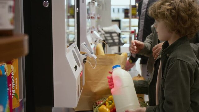 Pre-teen child with curly hair scanning plastic milk canister and giving it to unknown mother who packing groceries in paper bag at self-checkout