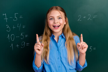 Smiling schoolgirl raising hands in a classroom with equations on a chalkboard, expressing learning...