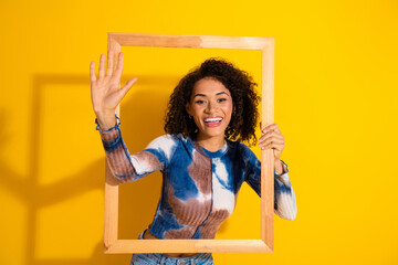 Happy young woman posing with a wooden frame against a bright yellow background, showcasing her...