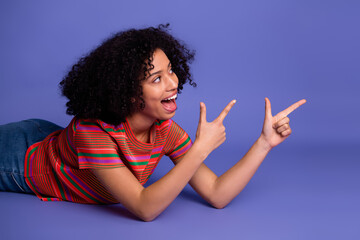 Young cheerful woman gesturing with pointed fingers, smiling while lying on violet backdrop in...