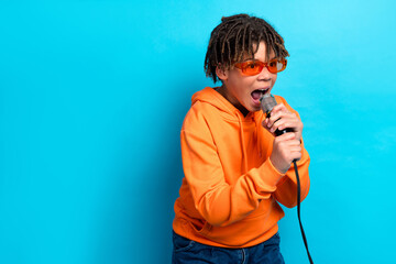 Energetic young boy karaoke singing with microphone in orange hoodie against vibrant blue background