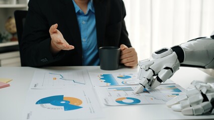 A human and a robot engage in a business meeting, discussing charts and data analysis, illustrating...