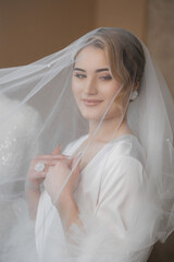 Bride in elegant wedding attire poses with a veil in a softly lit setting before ceremony