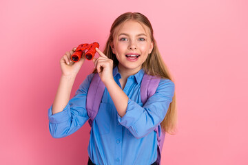 Blonde schoolgirl with purple backpack and red binoculars standing against a pink background...