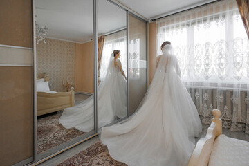 Bride in elegant wedding dress prepares in bright room with mirrors and curtains