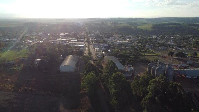 Drone ascending with the sun in the background and the city of Roncador below. The image has a greenish/orange hue due to the sunset, and in the city, a silo and warehouses can be seen.