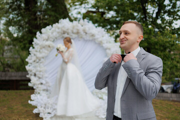 Groom adjusts suit before outdoor wedding ceremony with floral backdrop in springtime setting