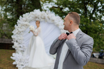 Groom adjusting bow tie while bride poses in elegant white wedding attire outdoors