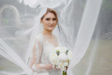 Bride in elegant white gown holds bouquet while surrounded by flowing veil on a rainy day