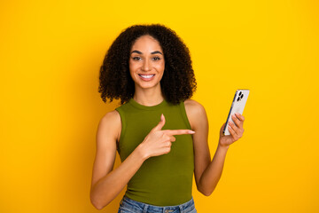 Young woman with curly hair holding a smartphone and pointing at it, in casual summer fashion...