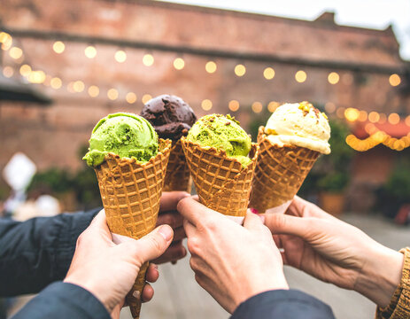 Four hands holding ice cream cones with diverse flavors, celebrating outdoors.