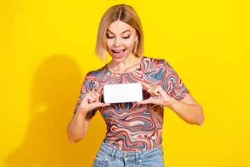 Cheerful young woman showing smartphone with excitement in colorful top against vibrant yellow...