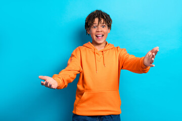 Happy teenager in orange hoodie gesturing openly on blue background, expressing joy and excitement...