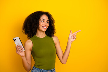 Smiling young woman with curly hair holding phone and pointing against yellow background in casual...