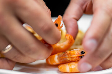 Close-up of human hands peeling cooked shrimp on a plate, showcasing vibrant orange seafood and...