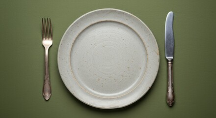 A top down view of a rustic light gray stoneware plate with an irregular edge Vintage silver fork and knife are positioned on either side The scene is set against a solid olive green background photor