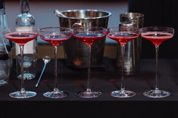 Row of five red cocktails in coupe glasses on black table with shaker, bottle and ice bucket. Indoor bar setting. Concepts: cocktail party, celebration, nightlife, alcohol menu, mixology,bar service. 