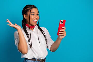 Young cheerful woman holding a red smartphone and interacting against a vibrant blue background,...