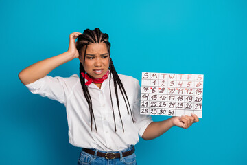 Young woman with braided hair holding a marked calendar on blue background, expressing confusion...