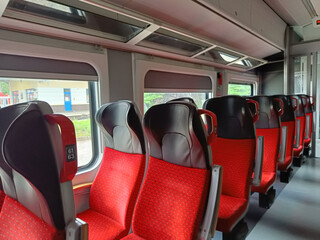 Fototapeta premium Row of empty red train seats next to the windows, with a station building visible outside. A calm and clean setting suitable for concepts like waiting, travel, or transition.