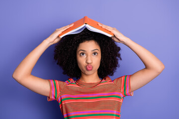 Young woman making a funny face with a book on her head, expressing creativity and humor in a casual and vibrant setting