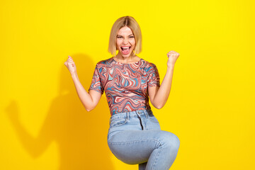 Excited young blonde woman raising fists in celebration, wearing colorful casual outfit against...