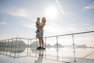 Caucasian woman holds her little son in her arms while standing on a glass bridge in the mountains. 