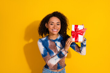Young female holding a beautifully wrapped gift box and smiling against a vibrant yellow...