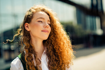 Confident young businesswoman with curly red hair standing outdoors in an urban setting on a sunny...