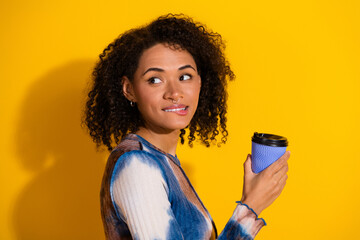 Charming young woman with curly hair smiling casually while enjoying a coffee in a stylish outfit...