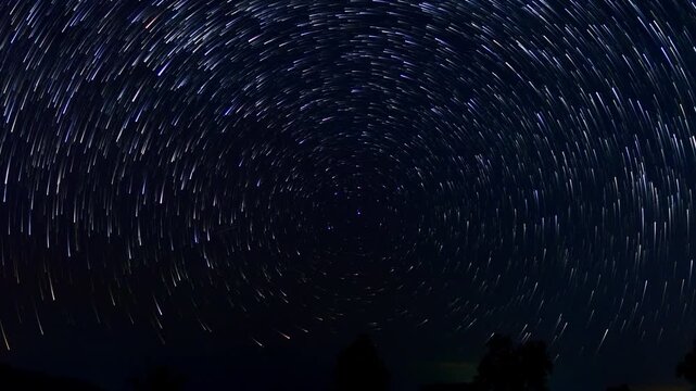 Time-lapse video showing star trails forming a half-circle across the night sky. The rotating stars create a mesmerizing pattern above a calm and dark landscape. 