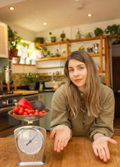 Light-skinned woman in olive shirt sitting at wooden kitchen table with mechanical scale full of red vegetables, looking at camera with open palms